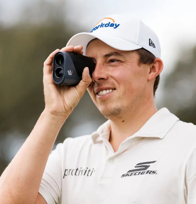 Image of a person wearing a white polo shirt and cap, holding a black rangefinder device up to their eye.